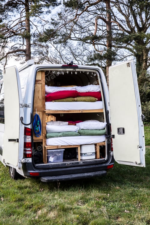 Preview: Rear view of camper van parked on a meadow, stacks of mattresses and bedding in back of vehicle.