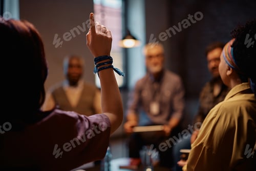 Preview: Rear view of woman with raised hand during group therapy at mental health center.