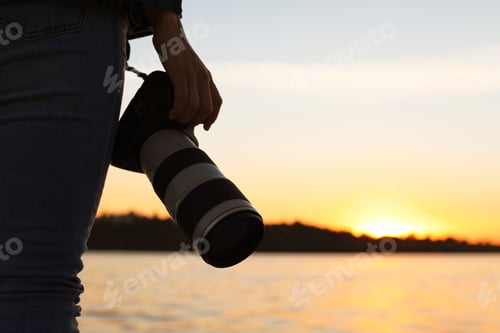 Preview: Female photographer with professional camera on riverside at sunset