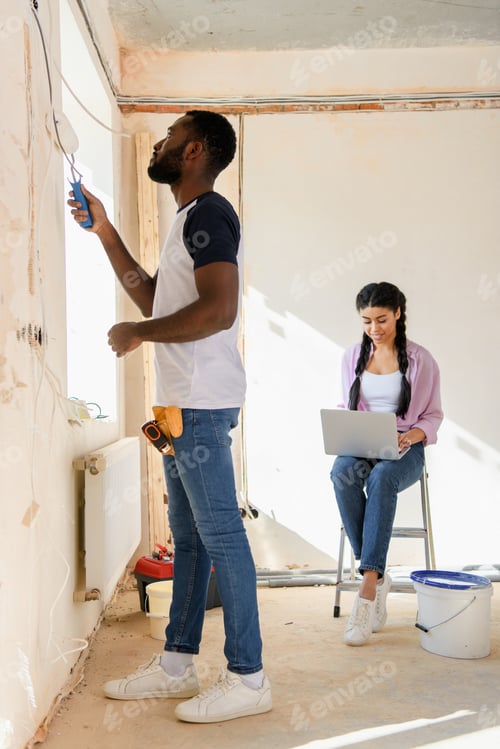 Preview: side view of african american man painting wall while his girlfriend using laptop behind during