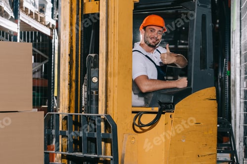 Preview: smiling warehouse worker showing thumb up and looking at camera while operating forklift loader