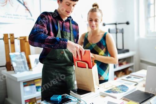 Preview: Young craftsman with customer, placing book into bag in arts bookshop