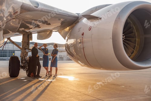Preview: Happy pilot and two pretty stewardesses standing together, leaning on an airplane