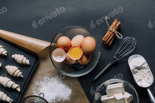 Preview: top view of ingredients, eggs in bowl and tray with dough for croissants on black table