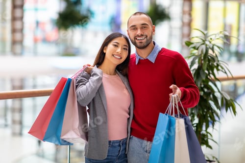 Preview: Portrait of young happy diverse couple posing with shopper bags, smiling at camera at huge city mall