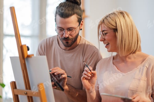 Preview: Precise creative couple at atelier painting together on easel.