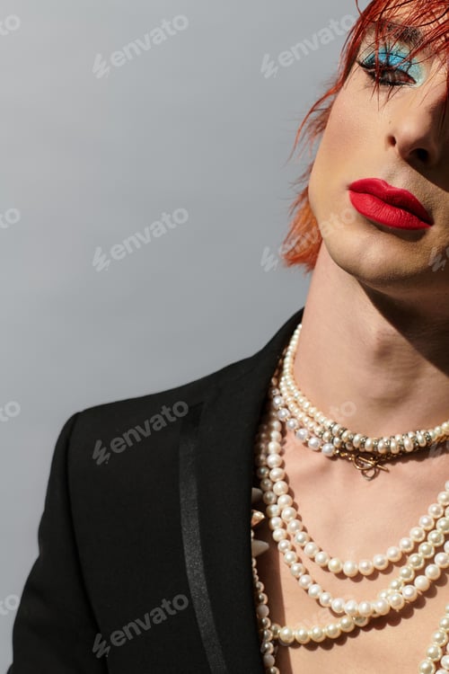 Preview: A young man showcases vibrant makeup and layered pearl necklaces against a gray backdrop.