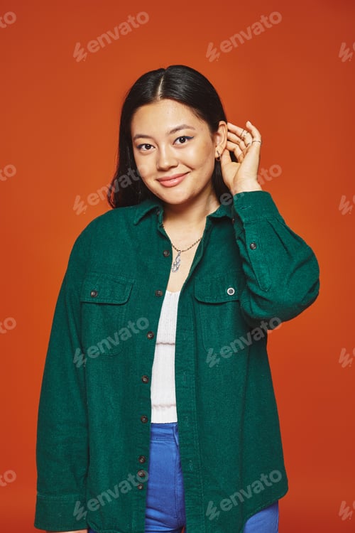 Preview: joyful young asian woman posing in vibrant outfit and adjusting brunette hair on orange background