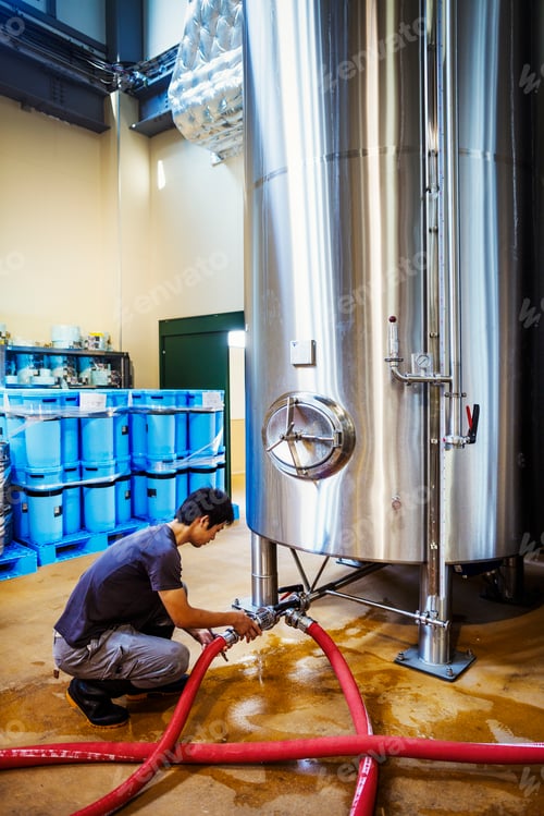 Preview: Man working in a brewery, connecting hoses to a metal beer tank.