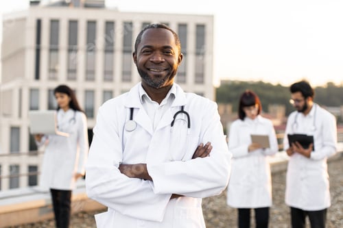 Preview: African doctor in white lab coat posing on camera with crossed hands and smiling