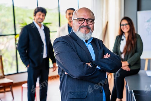 Preview: Confident oldIndian business leader standing with folded hands, colleagues out of focus in office