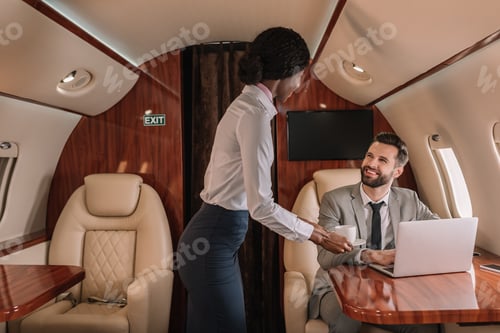 Preview: young african american stewardess giving cup of coffee to smiling businessman sitting at laptop in