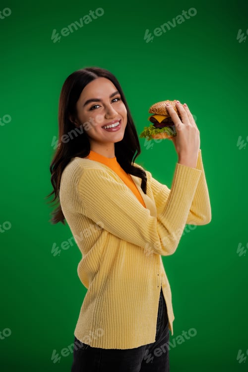 Preview: cheerful brunette woman looking at camera while holding tasty burger isolated on green