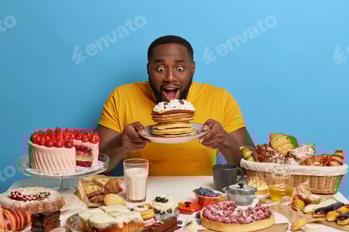 Preview: Dark skinned man tempted by delicious creamy pancakes, opens mouth, wears yellow t shirt, has sweet
