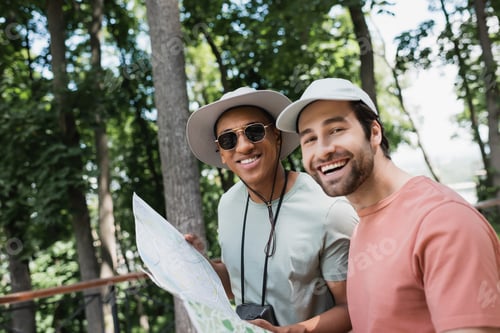 Preview: carefree interracial travelers holding map and smiling at camera in green park