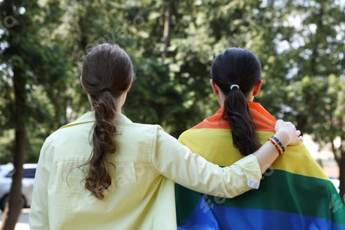 Preview: LGBT concept. Women in rainbow wristband with flag outdoors, back view