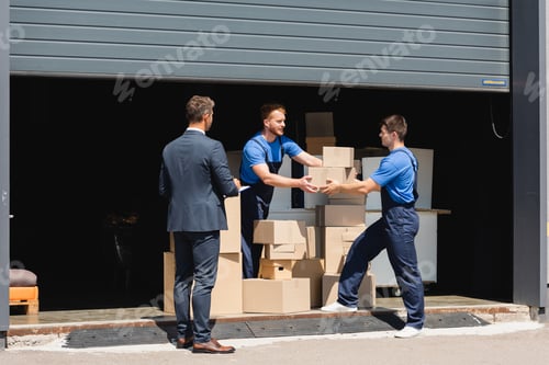 Preview: Businessman with clipboard standing near movers holding cardboard boxes in warehouse outdoors