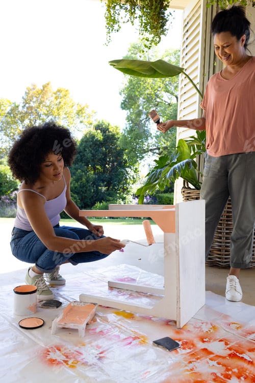 Preview: Mature biracial woman watching young biracial woman painting table at home for an upcycling project