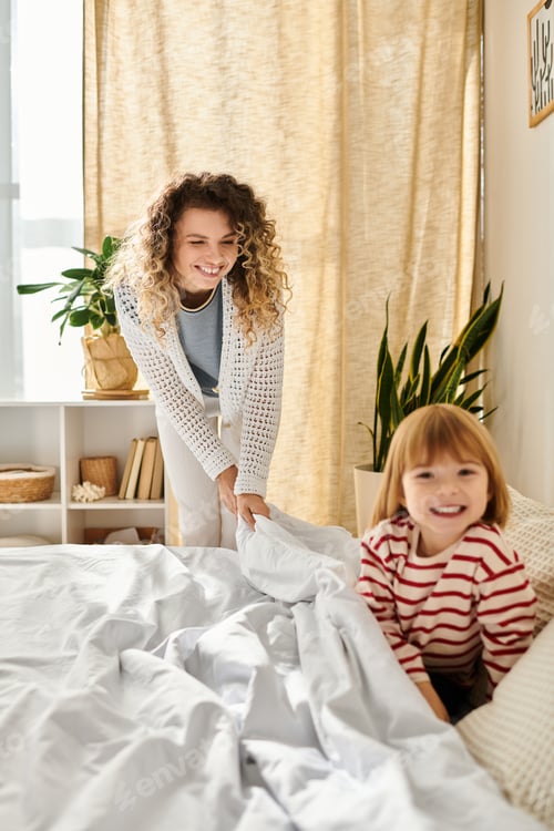 Preview: Curly-haired mother and daughter share laughter while making the bed in their cozy home.