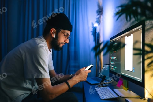 Preview: Young man mobile developer writes program code on a computer and check app in smartphone