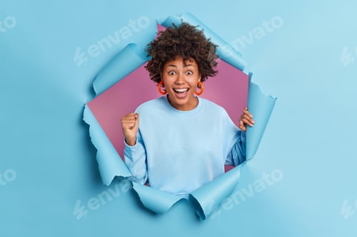 Preview: Excited dark skinned young woman with curly hair raises clenched fist and smiles broadly awaits for