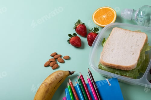 Preview: Sandwich in lunch box by fruits, almonds and color pencils with books on green background