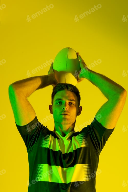 Preview: Portrait of caucasian male rugby player with rugby ball over yellow lighting