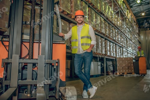Preview: Warehouse. Young Male Worker In Yellow Vest And Jeans Standing Near Shelves