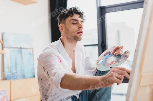 Preview: selective focus of handsome artist painting on canvas with paintbrush