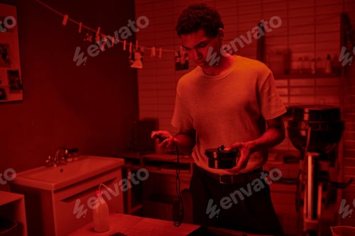 Preview: photographer in red-lit room, black man carefully handles film development with darkroom timer