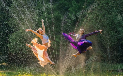 Preview: Two dancers in colorful costumes leap through a water spray against a lush green background