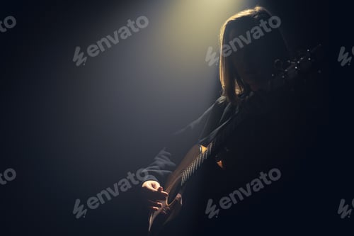Preview: A young woman with an acoustic guitar in the dark under a ray of light.