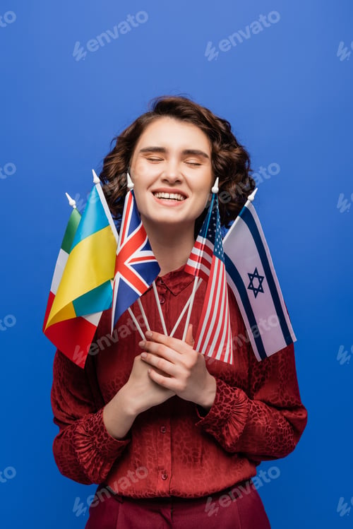 Preview: happy young woman holding international flags and smiling with closed eyes isolated on blue