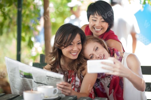 Preview: Three female friends posing for smartphone selfie at sidewalk cafe in city