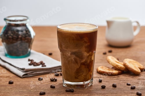 Preview: selective focus of ice coffee mixing with milk in glass near milk jug, cookies and coffee grains on