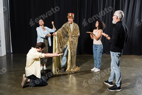 Preview: redhead man in king costume rehearsing role near multiethnic actors and art director in acting