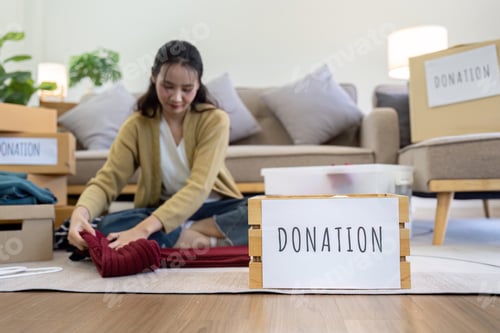 Preview: Asian young woman packing clothes at home, putting on stuff into donate box with second hand clothes