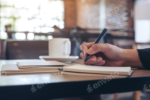 Preview: Closeup image of a hand writing on blank notebook with coffee cup on table in cafe