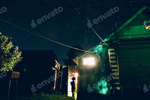Preview: Silhuette of kid in electric light outdoors near a house under a night star sky