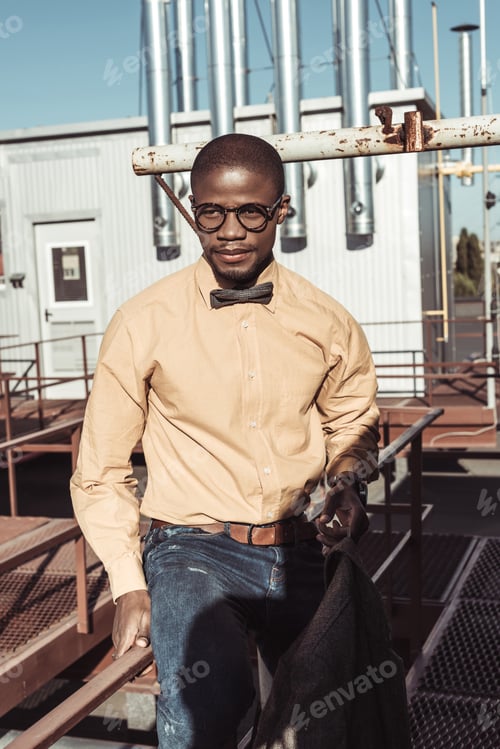 Preview: Stylish african american man wearing glasses and bowtie, sitting on guardrails and looking aside