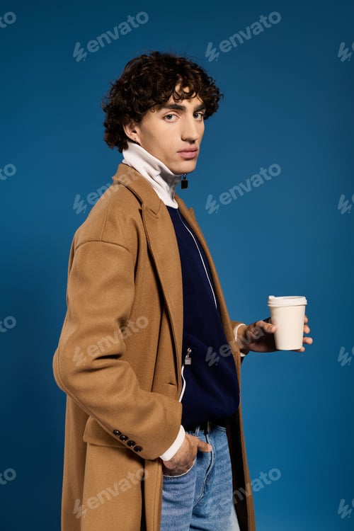 Preview: Stylish young man in fashionable coat enjoying coffee against a vibrant blue backdrop
