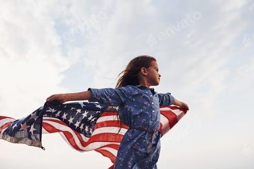 Preview: Patriotic female kid with American Flag in hands. Against cloudy sky