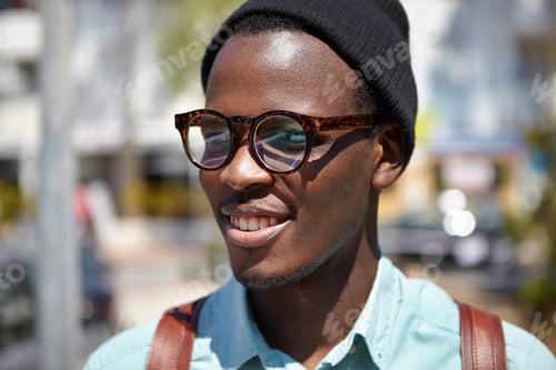 Preview: Close up summer portrait of handsome cheerful young black European student in trendy eyewear and hat