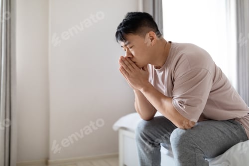 Preview: Side view frustrated asian man sitting on bed at home