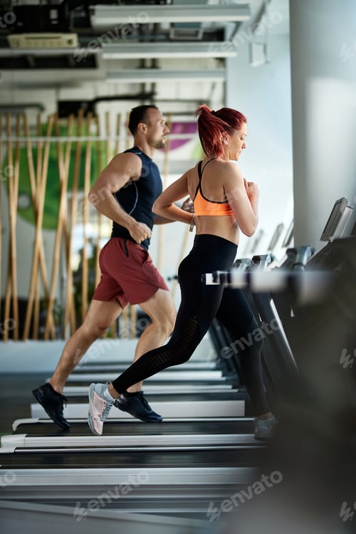 Preview: Athletic couple jogging on treadmills during sports training in a gym.