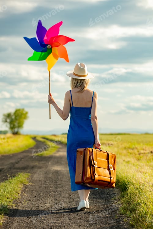 Preview: Girl in blue dress with suitcase and pinwheel on country road in summer.