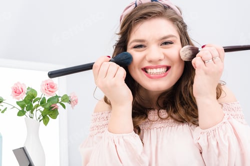 Preview: attractive young woman with makeup brushes in hands looking at camera at home
