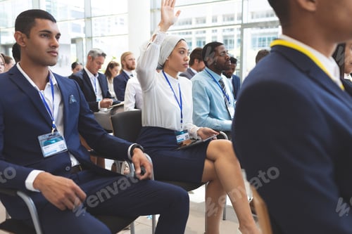 Preview: Side view of mixed race businesswoman raising hand in business seminar in office building