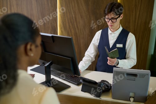 Preview: Young Asian man in uniform and eyeglasses checking personal data of guest