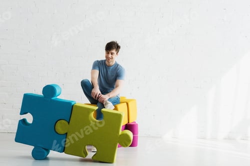 Preview: cheerful man looking at blue and green jigsaw pieces while sitting near brick wall